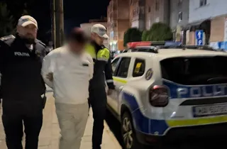 Un bărbat cu haine albe este condus de doi polițiști între un vehicul de serviciu alb și un trotuar noaptea.