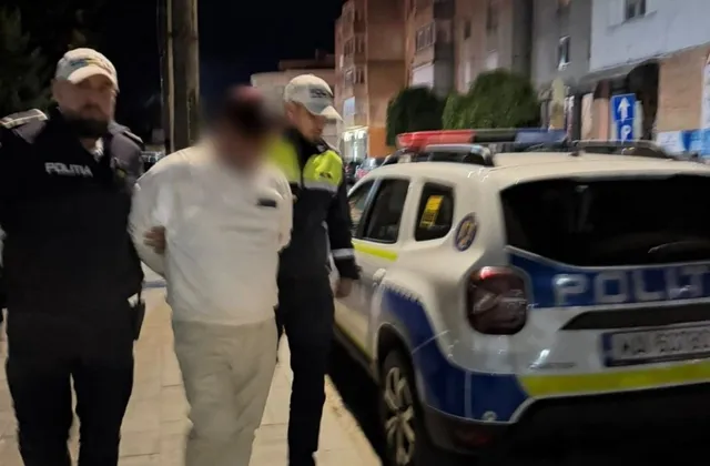 Un bărbat cu haine albe este condus de doi polițiști între un vehicul de serviciu alb și un trotuar noaptea.