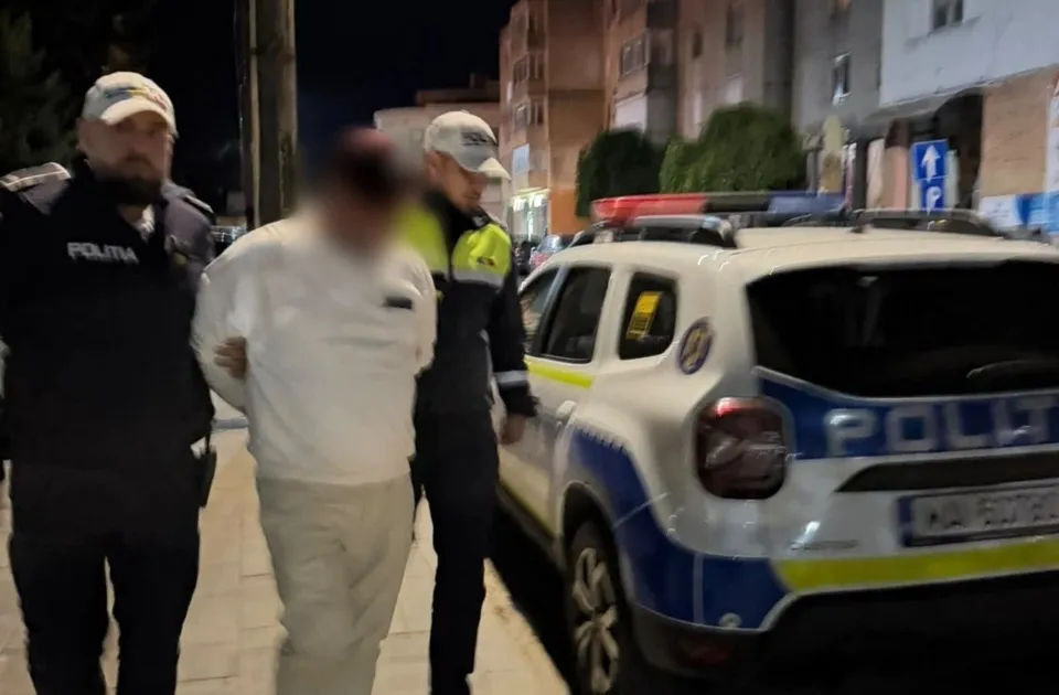 Un bărbat cu haine albe este condus de doi polițiști între un vehicul de serviciu alb și un trotuar noaptea.