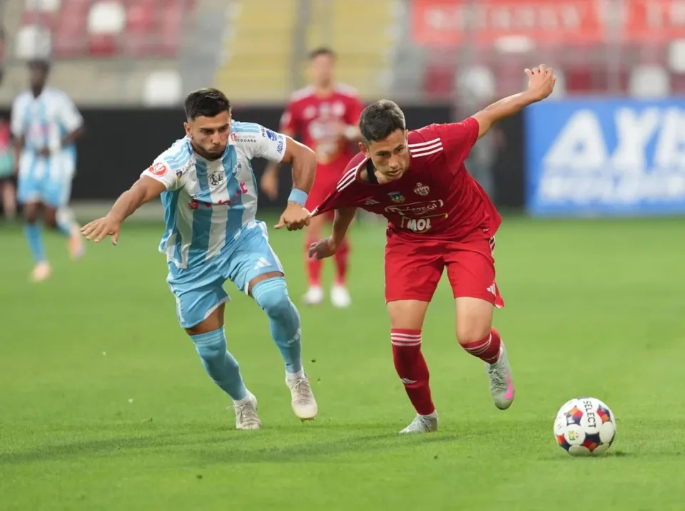 Un jucător de fotbal în echipament roșu conduce mingea pe teren, fiind urmărit de un alt jucător îmbrăcat în echipament albastru și alb.