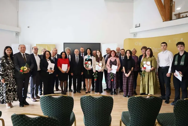 Un grup de adulți și tineri stă în rând, purtând haine formale, într-o încăpere cu podea de lemn și scaune verzi în prim-plan.