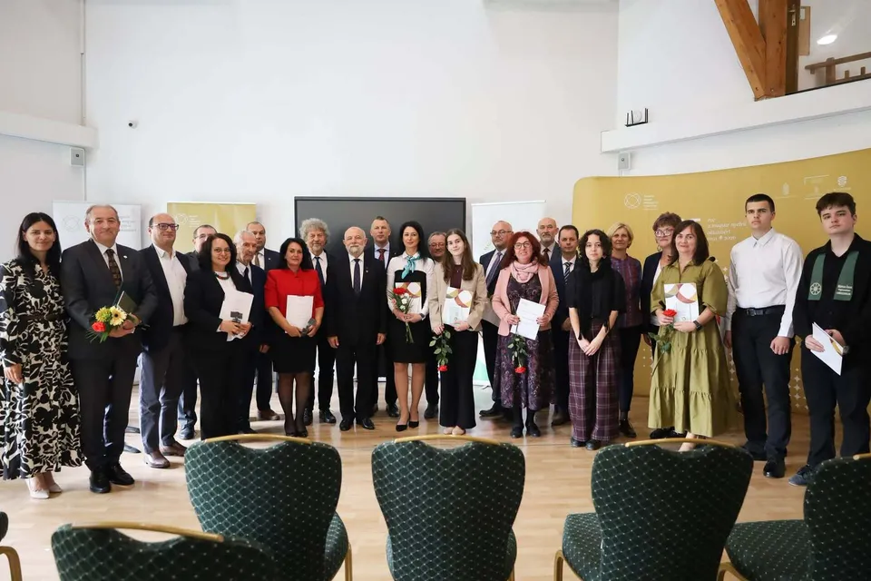 Un grup de adulți și tineri stă în rând, purtând haine formale, într-o încăpere cu podea de lemn și scaune verzi în prim-plan.