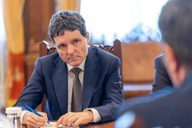 Un bărbat cu părul închis la culoare, purtând sacou albastru și cravată închisă, stă la o masă de lemn ținând un pix albastru.