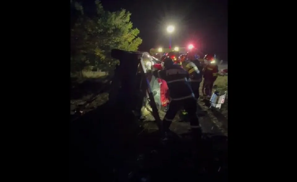 Mai mulți pompieri în uniforme de lucru întunecate lucrează lângă un autoturism răsturnat pe marginea unei drumuri sub lumină artificială noaptea.