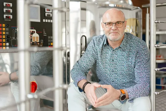 Un bărbat cu ochelari și cămașă cu modele mici stă sprijinit de o structură metalică lângă un echipament industrial de oțel inoxidabil.