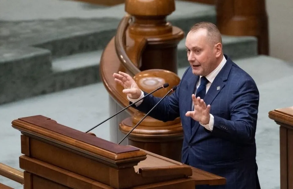 Un bărbat în costum la un trib cu microfoane, într-un interior cu balustradă din lemn.