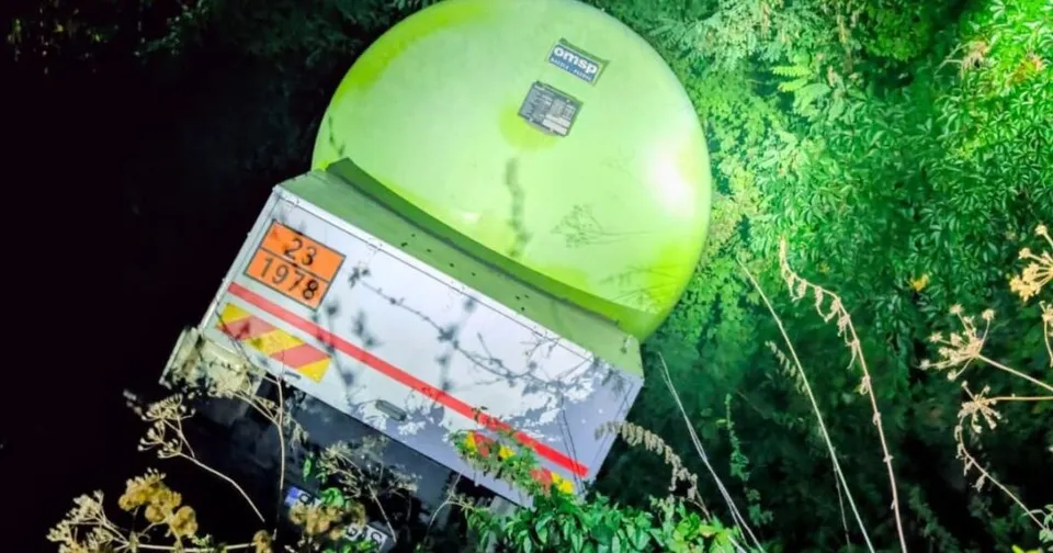 O autocisternă cu rezervor verde și caroserie albă, cu dungi roșii, este înclinată printre vegetație și crengi.