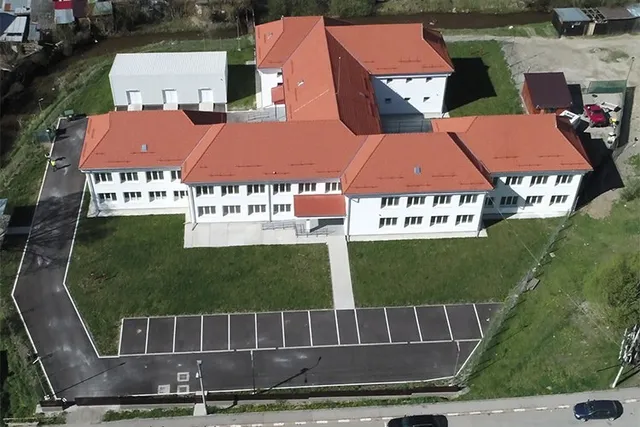 O vedere aeriană prezintă un complex de clădiri albe cu acoperișuri roșii, înconjurat de o curte cu iarbă verde și un parcare asfaltat.