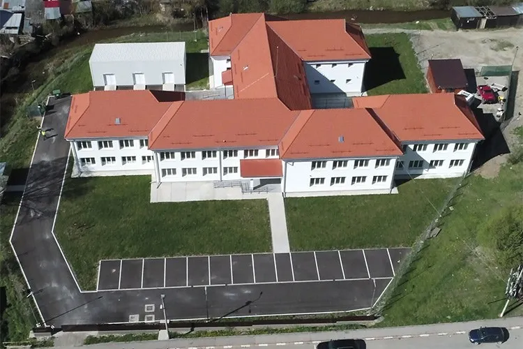 O vedere aeriană prezintă un complex de clădiri albe cu acoperișuri roșii, înconjurat de o curte cu iarbă verde și un parcare asfaltat.