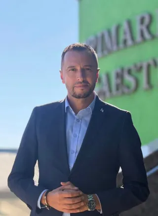 Un bărbat cu părul scurt, purtând un sacou închis la culoare și o cămașă deschisă, stă cu mâinile împreunate în față.