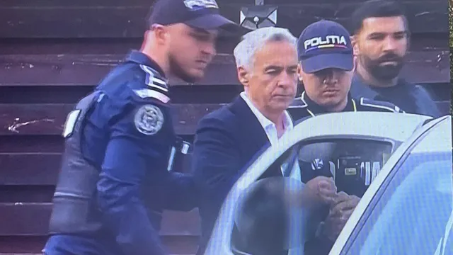 Un bărbat cu părul cărunt și sacou închis este condus de polițiști în uniformă lângă o mașină albă.