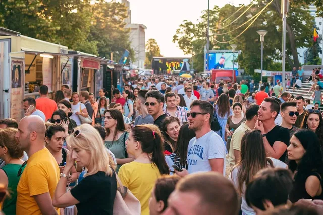 O mulțime de oameni stă în picioare într-o zonă deschisă lângă containere de tip food truck, sub copaci și cerul de seară.