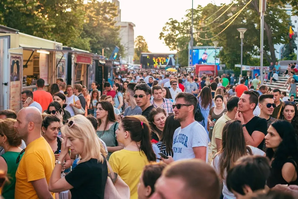 O mulțime de oameni stă în picioare într-o zonă deschisă lângă containere de tip food truck, sub copaci și cerul de seară.