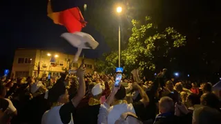 O mulțime de oameni stă în noapte, ridicând telefoane mobile și un steag cu culorile roșu, alb și albastru sub un stâlp de iluminat stradal.