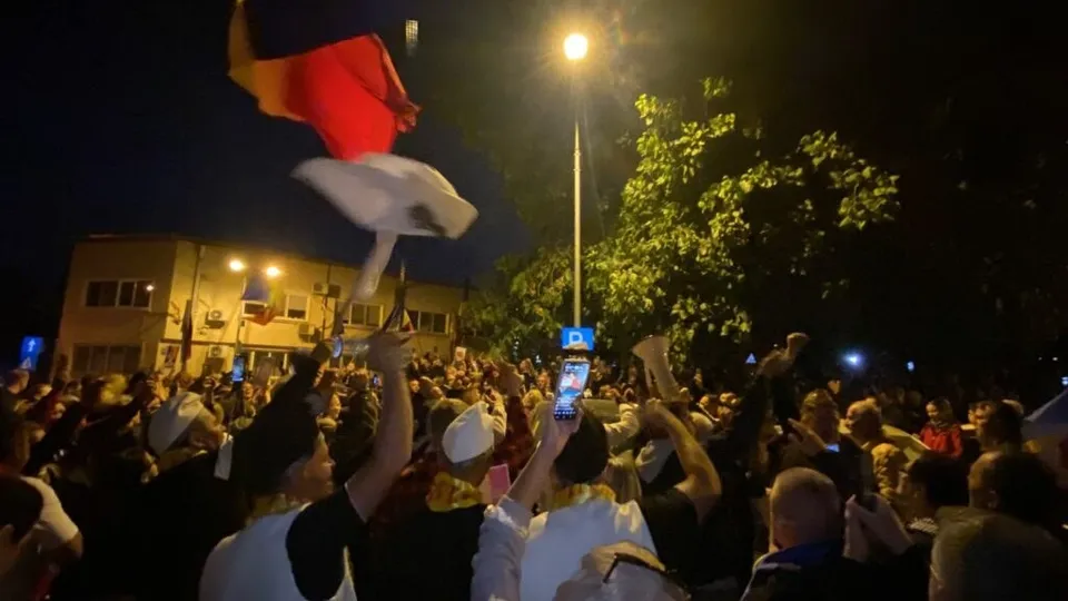O mulțime de oameni stă în noapte, ridicând telefoane mobile și un steag cu culorile roșu, alb și albastru sub un stâlp de iluminat stradal.
