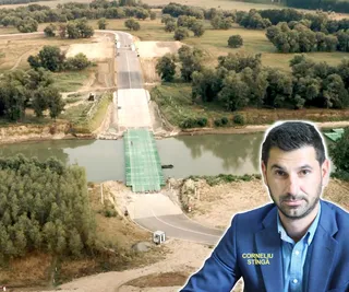O fotografie de sus arată o porțiune de drum care traversează un râu prin intermediul unui pod cu suprafață verde, lângă o zonă împădurită.