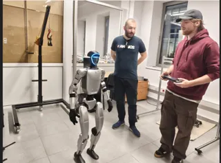 Un robot umanoid argintiu stă în picioare într-o încăpere lângă doi bărbați, unul purtând un hanorac roșu și celălalt un tricou închis la culoare.
