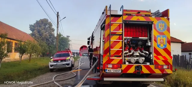 Un autospecial de pompieri cu portbagajul deschis este parcat pe un drum lângă un autoturism alb și o furtun de incendiu întins pe sol.