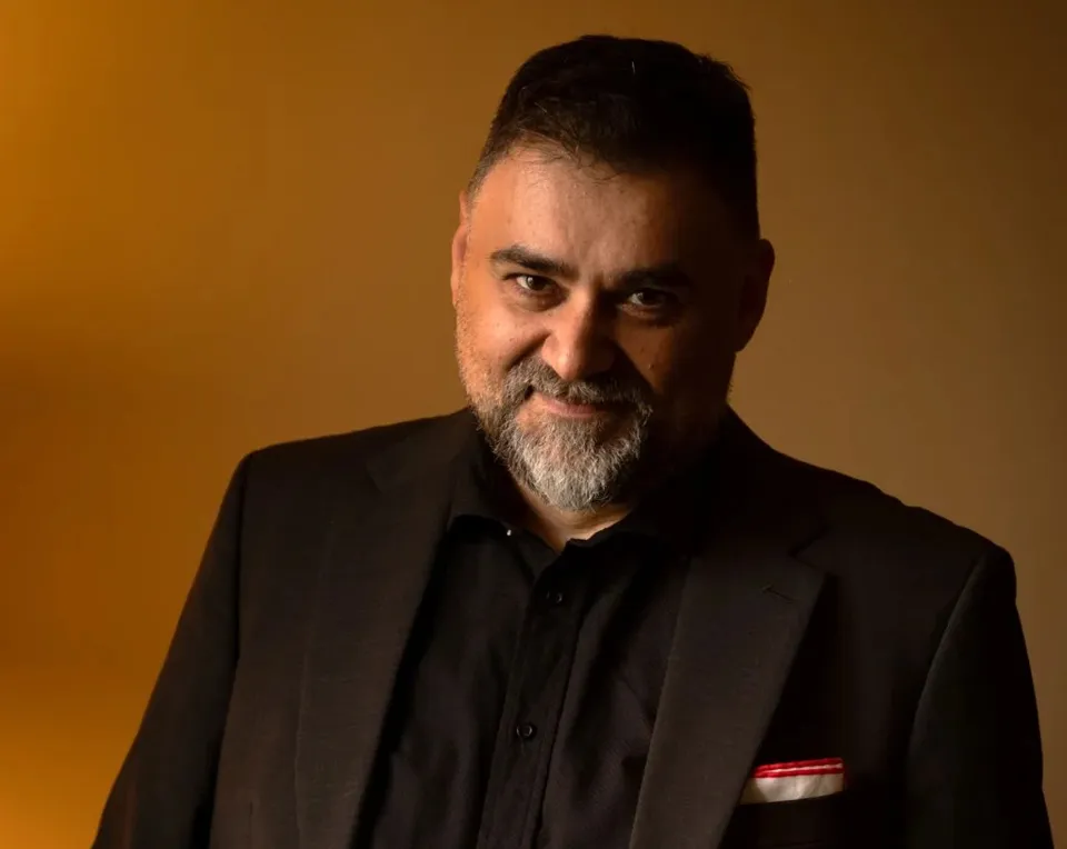 Un bărbat cu păr și barbă grizonante, îmbrăcat într-un costum negru, privește spre cameră pe un fundal maroniu.