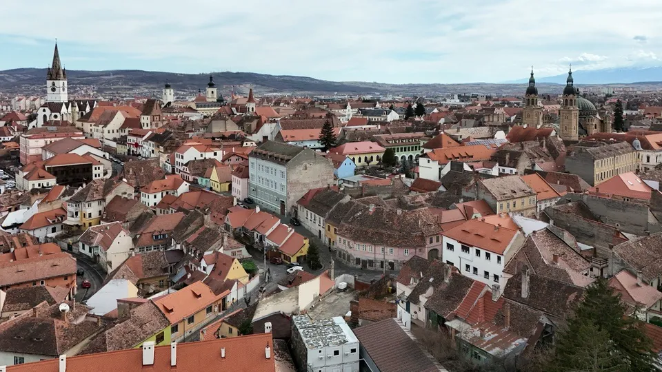 O vedere panoramică de la înălțime asupra unui oraș cu numeroase case cu acoperișuri de culoare roșie și câteva turnuri de biserică sub un cer înnorat.