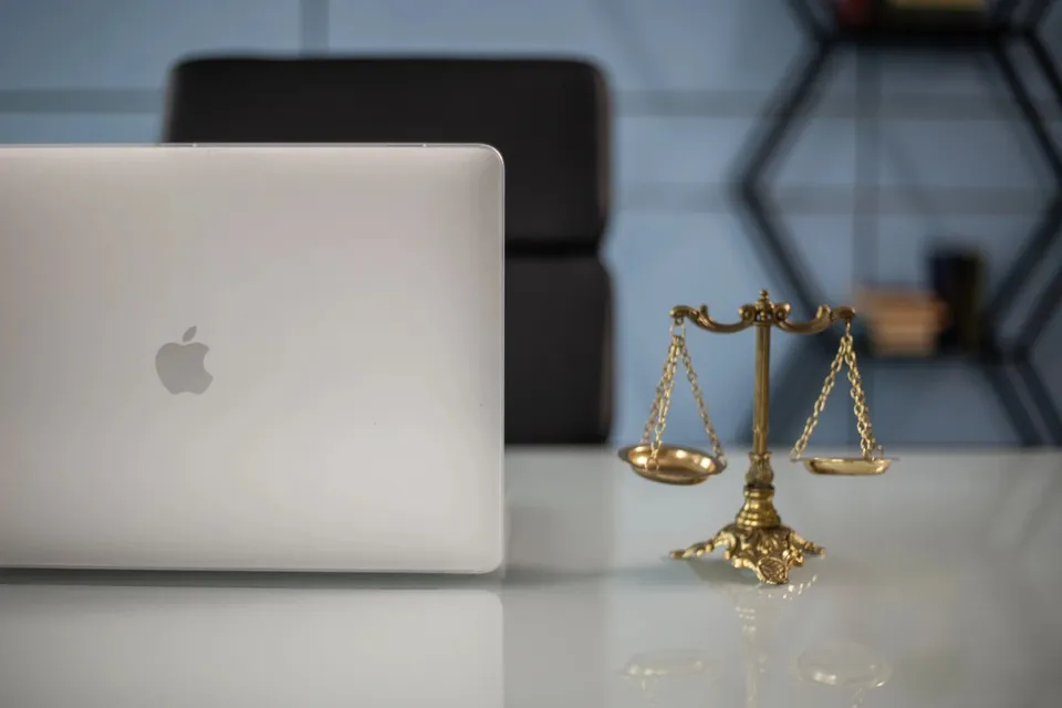 O balanță de metal aurie stă pe o suprafață albă lângă un laptop închis într-un decor cu fundal albastru.
