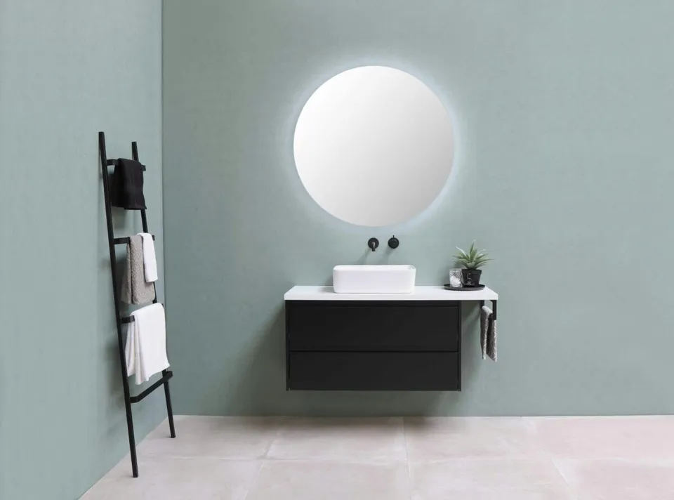 Un lavoar alb este așezat pe un mobilier suspendat negru sub o oglindă circulară iluminată, lângă un suport vertical pentru prosoape.