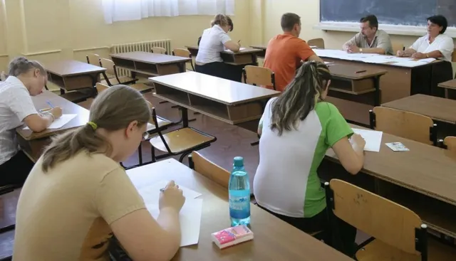 Oameni așezați la mese individuale în interiorul unei săli, scriind pe foi de hârtie albe.