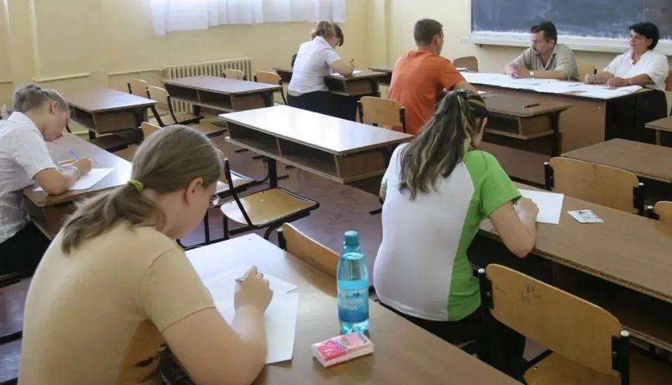 Oameni așezați la mese individuale în interiorul unei săli, scriind pe foi de hârtie albe.