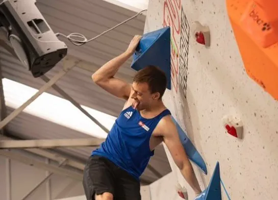 Un bărbat cu o cămașă albastră de sport și pantaloni negri se prinde de elemente de escaladă pe un perete alb.