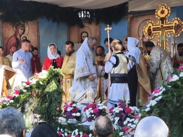 Câțiva bărbați îmbrăcați în haine liturgice albe și aurii stau în fața unor picturi religioase, înconjurați de flori colorate și ramuri verzi.