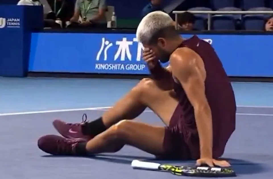 Un bărbat cu părul scurt și deschis la culoare stă așezat pe un teren de tenis, având capul aplecat și mâna pe față.