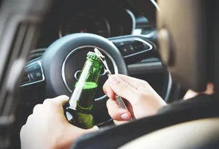 O persoană ține o sticlă de bere verde și un deschizător de sticle în interiorul unui autoturism, lângă volanul mașinii.