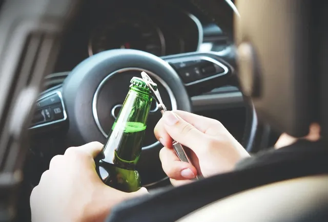 O persoană ține o sticlă de bere verde și un deschizător de sticle în interiorul unui autoturism, lângă volanul mașinii.