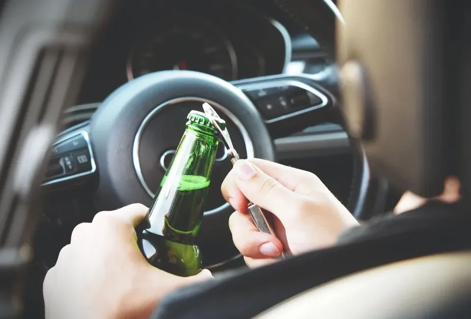 O persoană ține o sticlă de bere verde și un deschizător de sticle în interiorul unui autoturism, lângă volanul mașinii.
