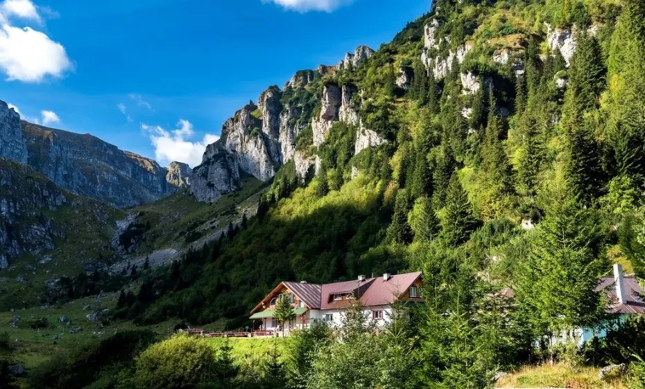 O clădire cu acoperiș roșu este situată la poalele unui munte abrupt acoperit de pădure de conifere sub un cer albastru cu nori albi.