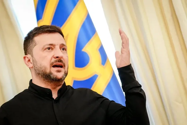 Un bărbat cu barbă și cămașă neagră stă în fața unui steag albastru cu elemente galbene, ridicând mâna stângă.