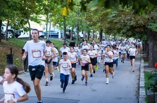 Un grup mare de oameni, inclusiv copii și adulți, aleargă pe un drum asfaltat înconjurat de copaci verzi.