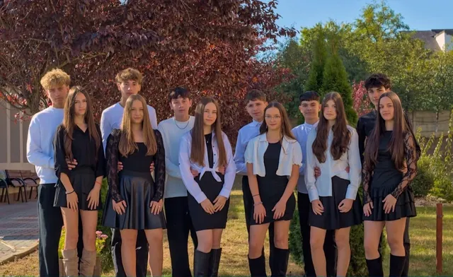 Un grup de tineri stă în rând, îmbrăcați în haine negre și albe, în fața unor copaci și a unei grădini verzi sub cerul senin.