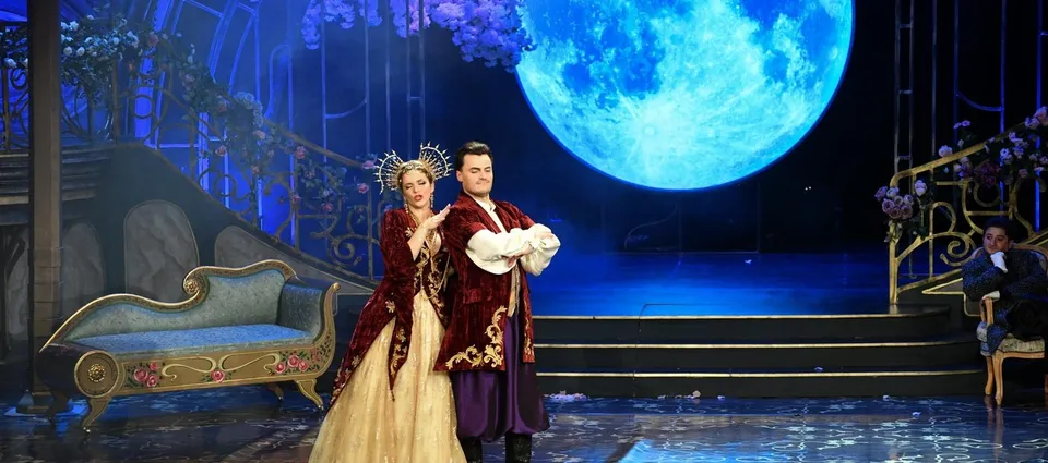 O femeie în rochie lungă și un bărbat în costum teatral stau pe o scenă decorată cu o lună mare și flori.