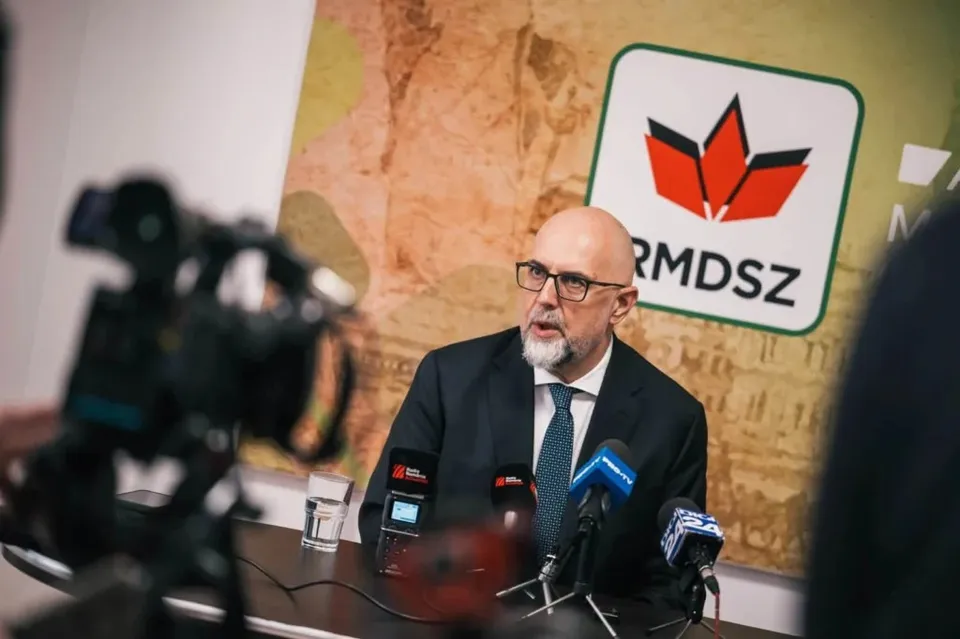 Un bărbat cu ochelari și barbă căruntie, purtând costum închis și cravată, stă la un microfon în fața unui panou cu un logo roșu și verde.