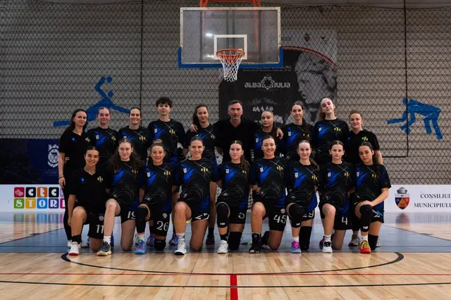 Un grup de femei îmbrăcate în echipamente sportive negre și albastre pozează într-o sală de sport cu un coș de baschet în fundal.