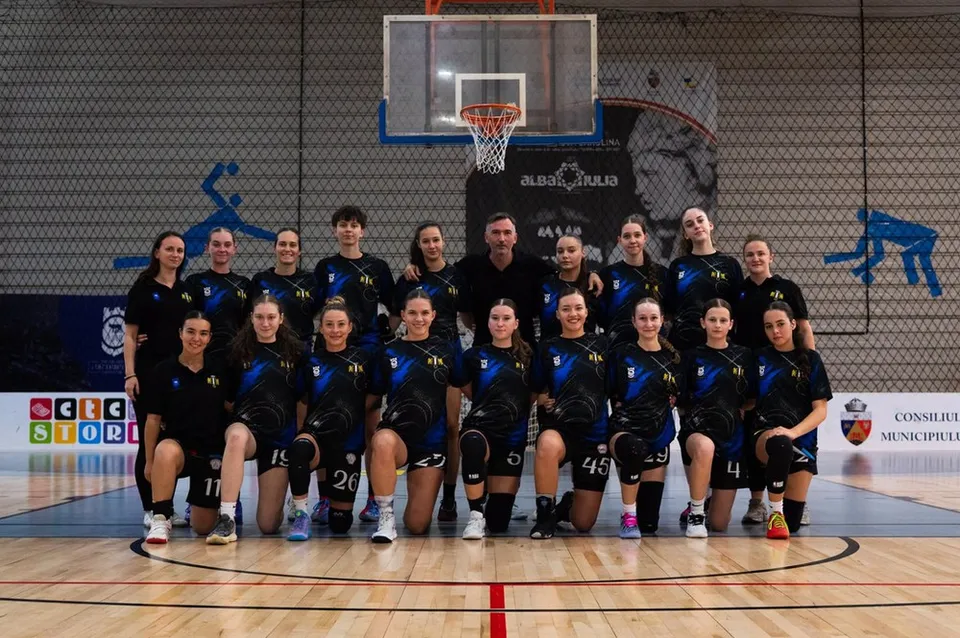 Un grup de femei îmbrăcate în echipamente sportive negre și albastre pozează într-o sală de sport cu un coș de baschet în fundal.