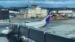 Un avion de pasageri este parcat pe o pistă de aeroport lângă o punte de îmbarcare acoperită, având un terminal în fundal.