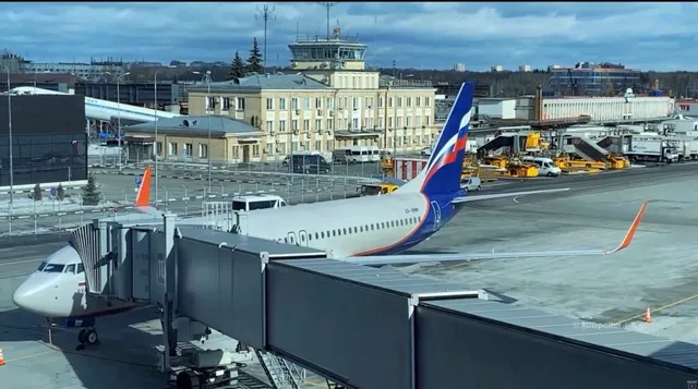 Un avion de pasageri este parcat pe o pistă de aeroport lângă o punte de îmbarcare acoperită, având un terminal în fundal.