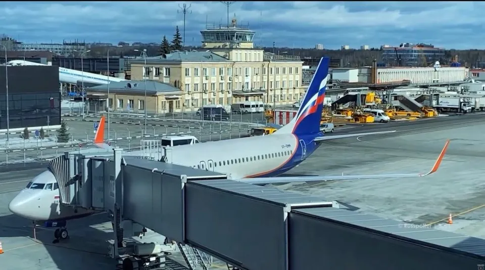 Un avion alb cu detalii albastre și roșii este parcat lângă o punte de îmbarcare într-un aeroport cu clădiri și vehicule în fundal.
