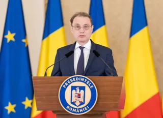 Un bărbat cu ochelari, purtând costum închis la culoare și cravată, stă în spatele unui podium cu un emblema circulară în fața unor steaguri.