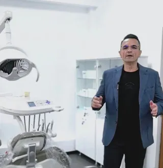 Un bărbat cu ochelari și sacou albastru stă într-o cameră albă lângă un scaun stomatologic și un dulap cu rafturi albe.