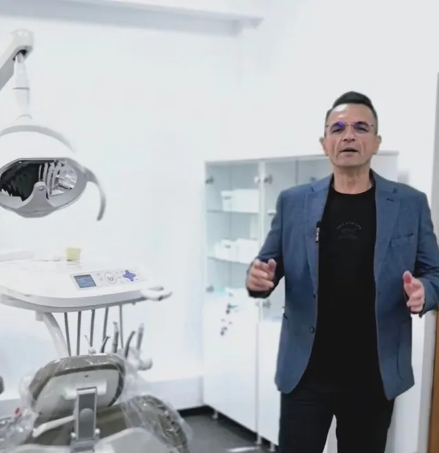 Un bărbat cu ochelari și sacou albastru stă într-o cameră albă lângă un scaun stomatologic și un dulap cu rafturi albe.