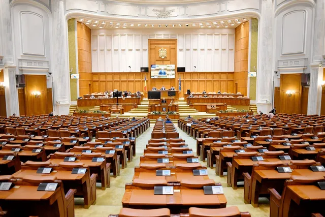 O sală de parlament cu numeroase birouri de lemn dispuse în rânduri și un podium central cu ecrane și panouri de lemn în fundal.