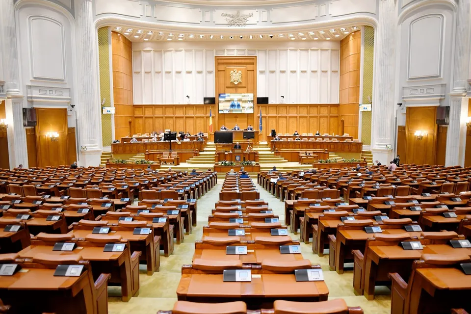O sală de parlament cu numeroase birouri de lemn dispuse în rânduri și un podium central cu ecrane și panouri de lemn în fundal.